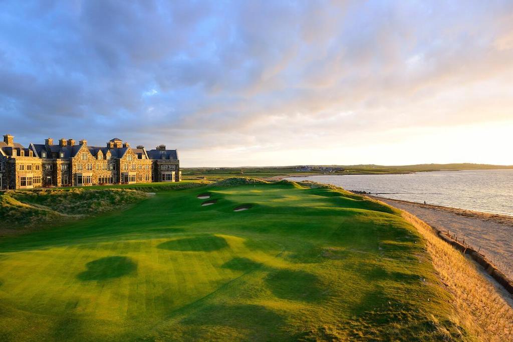 The coastal fairway at Doonbeg, County Clare, with a grand stone lodge beside the sea, lit by warm evening sunlight.