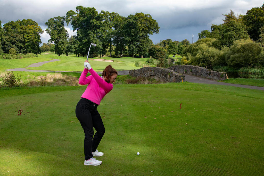 A woman swings her club on a green fairway at The K Club in County Kildare, with a stone bridge and trees behind her.