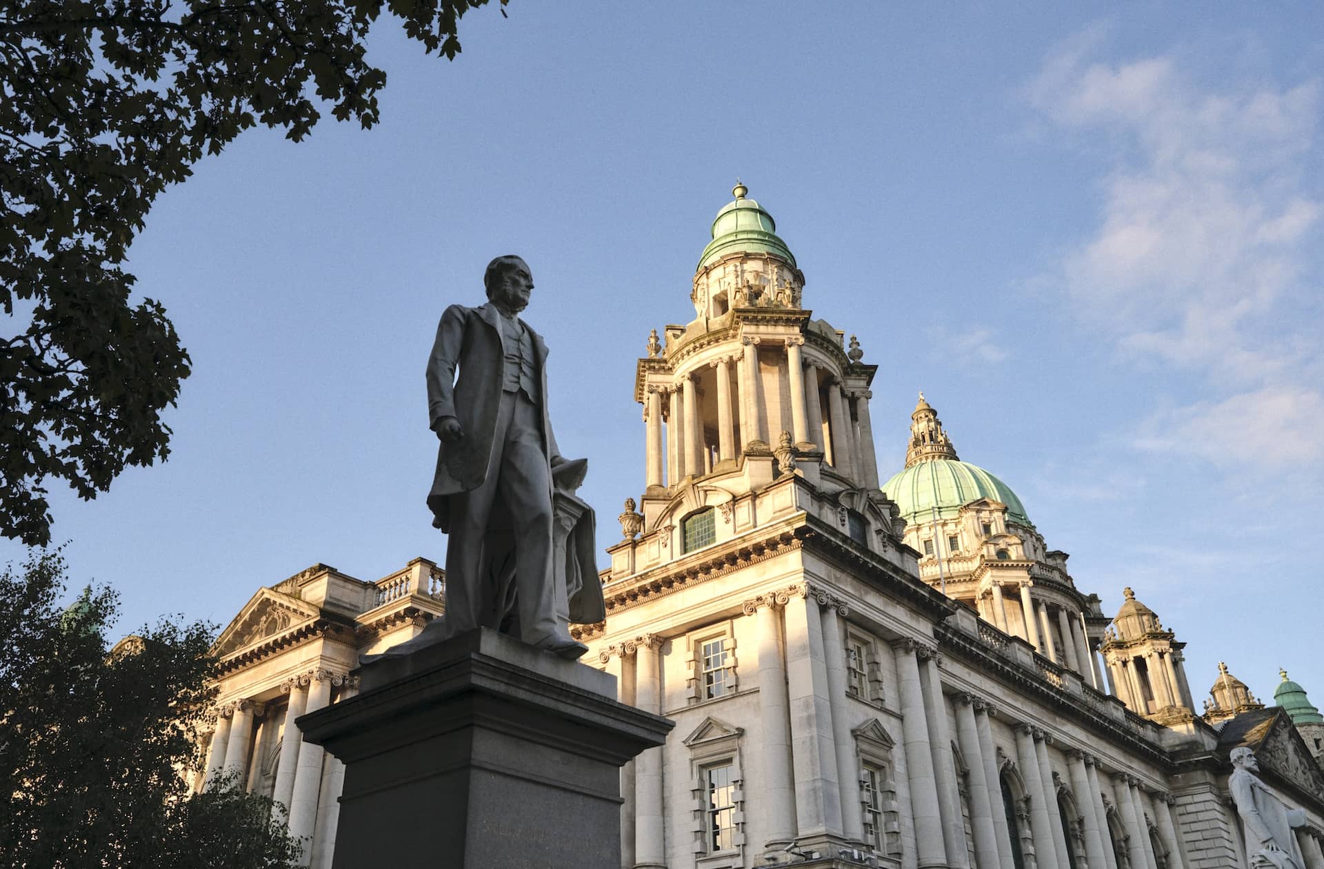 Eine Statue steht vor den prächtigen Kuppeln und der kunstvollen Fassade des Rathauses von Belfast unter einem klaren Abendhimmel.