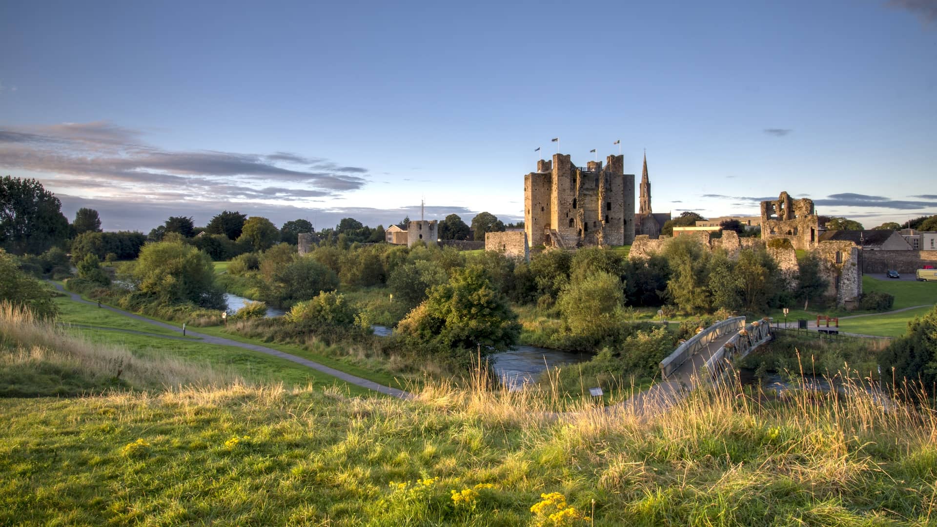 Trim Castle rises above the River Boyne at sunset, surrounded by green parkland and historic ruins in County Meath.