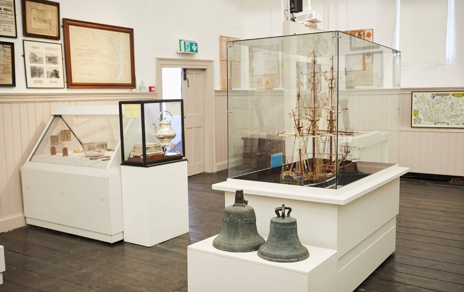Exhibit at Kinsale Museum with ship model in glass case, silver artifacts, and bells displayed on white stands.