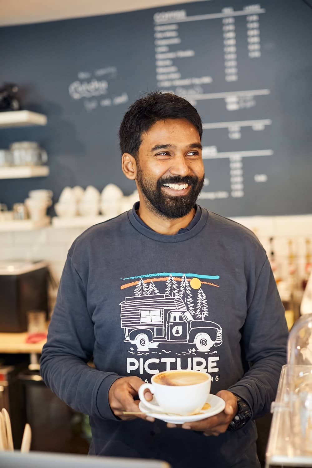 Smiling man holding a cappuccino at The Post House café, standing in front of a chalkboard menu.