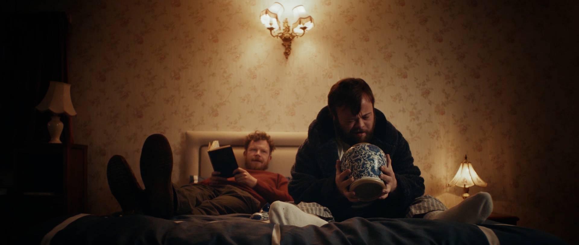 Zwei Männer in einem warm beleuchteten Schlafzimmer, einer liest im Bett, der andere sitzt und hält eine große blau-weiße Vase.