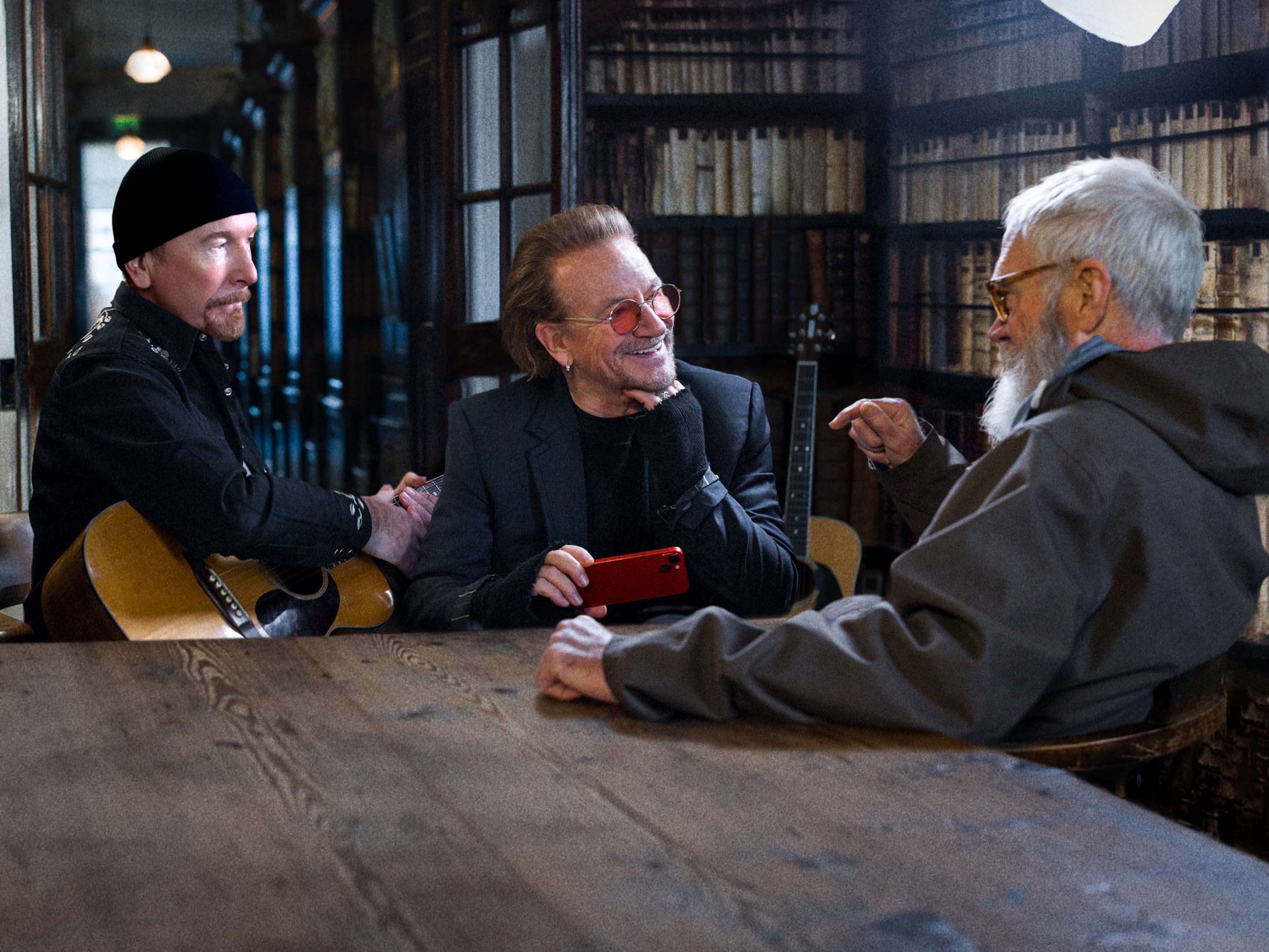 Bono und The Edge sitzen mit einem Mönch in einer Bibliothek, hinter ihnen stehen Gitarren, während der Dreharbeiten für A Sort of Homecoming.