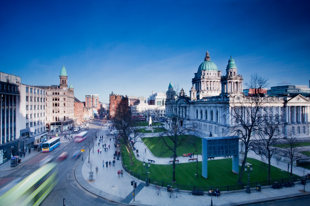 Blick auf das Rathaus von Belfast und die umliegenden Straßen an einem klaren Tag, mit Menschen, die spazieren gehen und vorbeirasendem Verkehr.