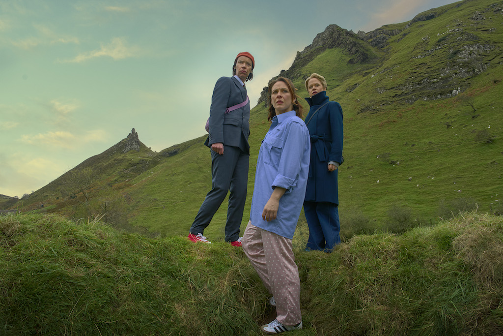 Still from How to get to Heaven from Belfast, showing three women on a hillside, looking off into the distance, with green mountains and a cloudy sky.