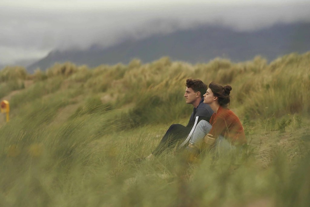Young couple sit in sandy dunes, looking out towards dark hills in the distance on Streedagh Beach, County Sligo.