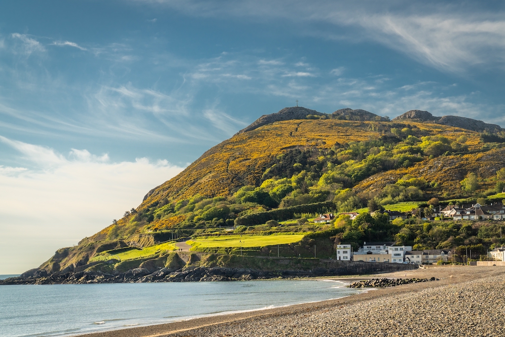 Sandy beach below a sunlit Bray Head with houses and trees beside a calm bay in Bray, County Wicklow.