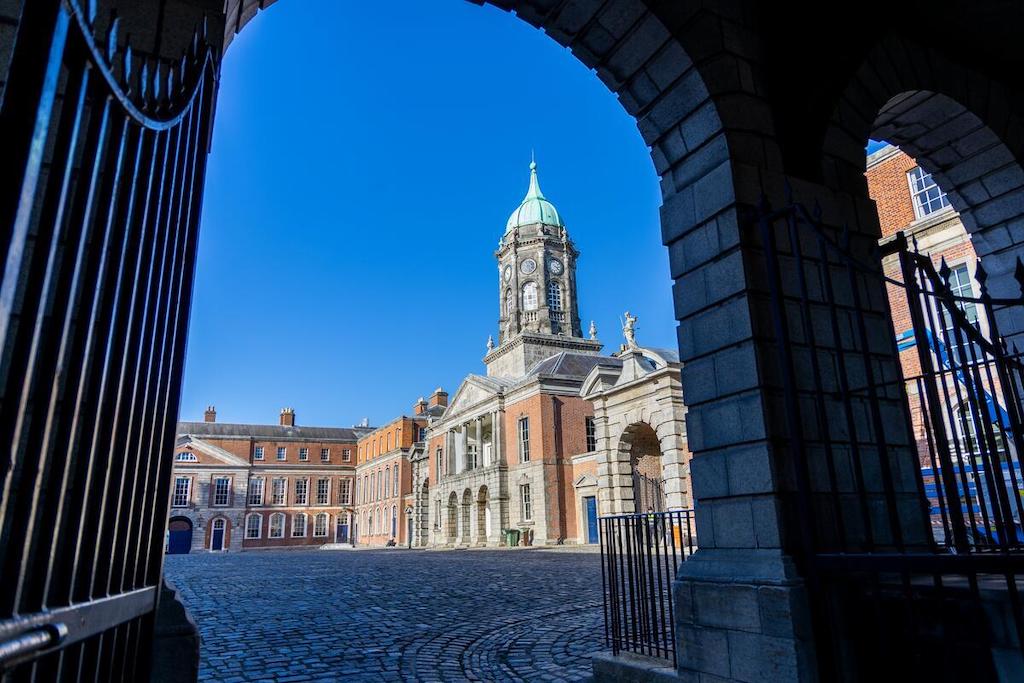 Dublin Castle courtyard framed by an archway, with the clock tower under a blue sky.
