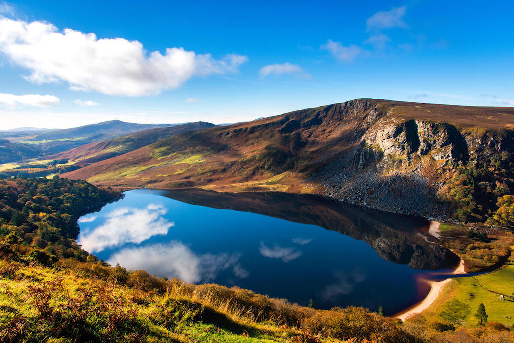 Panoramic view of Lough Tay in County Wicklow reflecting clouds, with a steep rocky cliff on the shore.