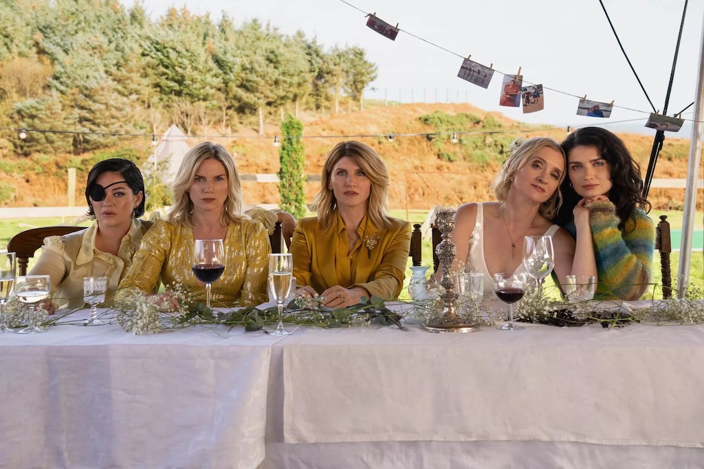 Five women from Bad Sisters sit at a long outdoor table with wine glasses, staring ahead in silence.