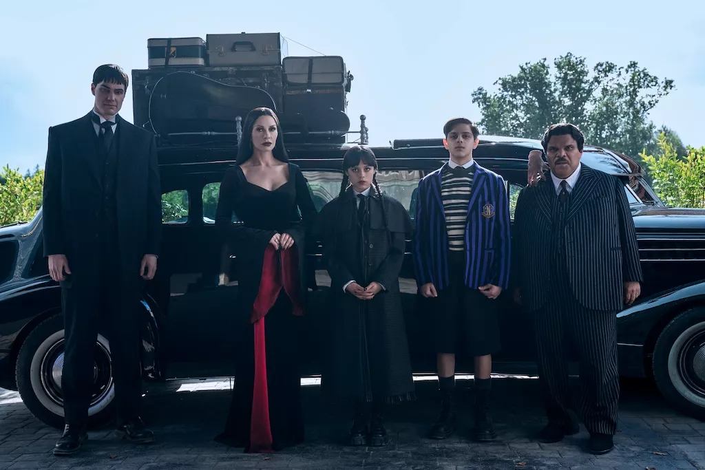 The Addams family stand in front of a black car with luggage stacked on the roof in Wednesday.