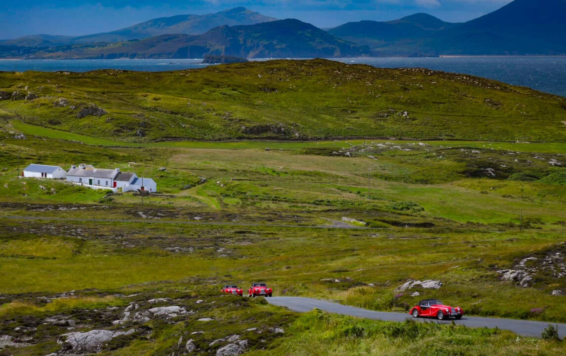 Red classic cars driving along a winding rural road past white cottages with mountains and sea views on the Wild Atlantic Way.