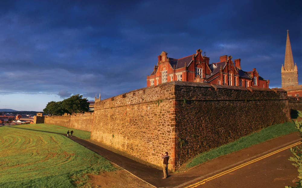 People walk along the 17th-century city walls of Derry~Londonderry with red-brick buildings in the golden light.