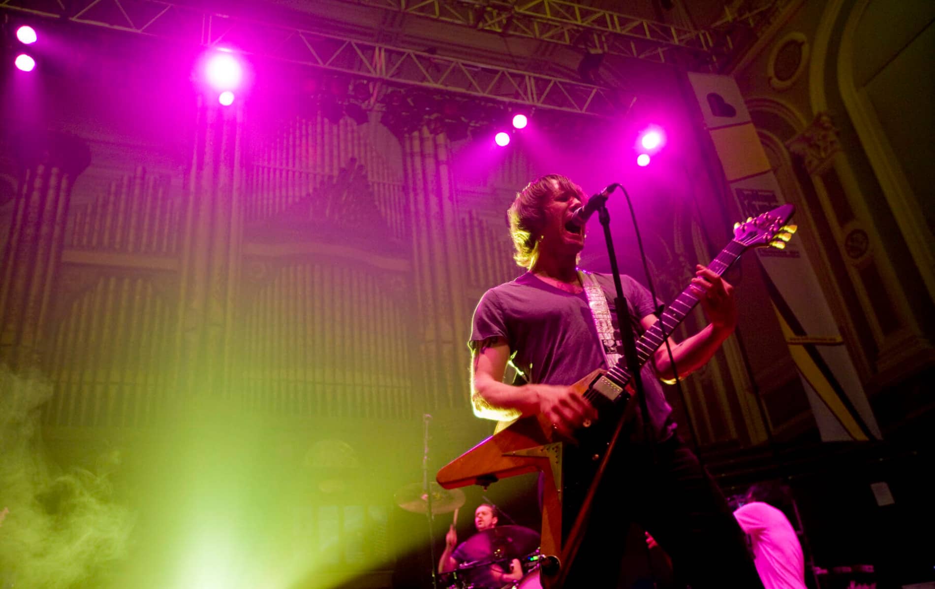 Rockmuzikant die zingt en een elektrische gitaar bespeelt onder paars en geel podiumlicht in de historische Ulster Hall in Belfast.
