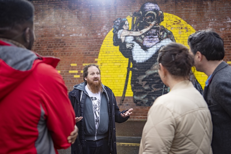 Una guida parla ai visitatori accanto a un grande murale su un muro di mattoni durante il Seedhead Arts Street Art Walking Tour a Belfast.