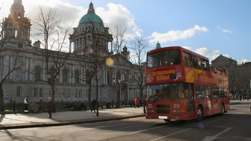 Un bus turistico rosso passa davanti al Belfast City Hall in una giornata soleggiata.