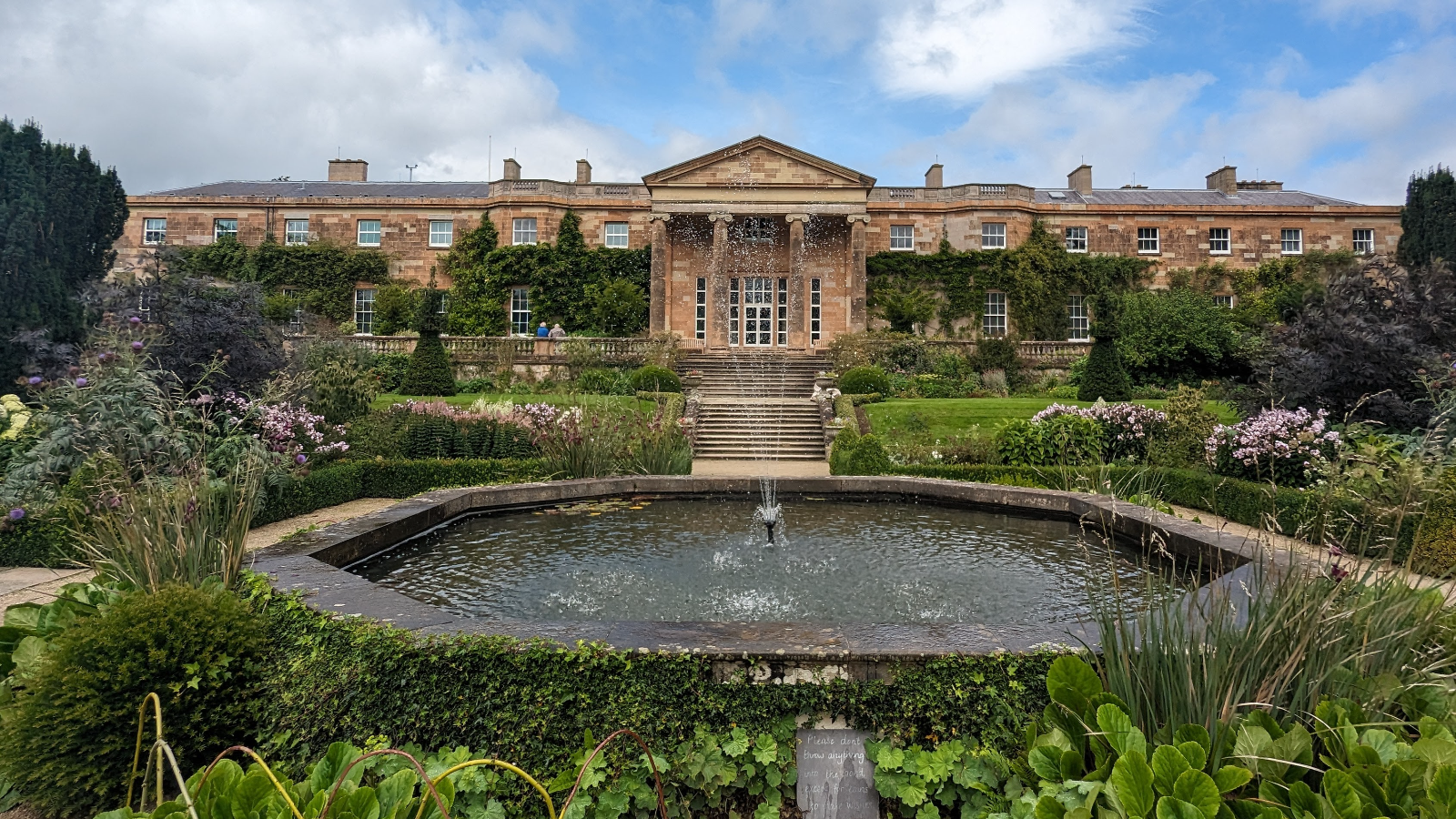 Il castello di Hillsborough, nella contea di Down: una sontuosa residenza storica che si affaccia su giardini formali e su uno stagno circolare con una fontana, in una giornata soleggiata.