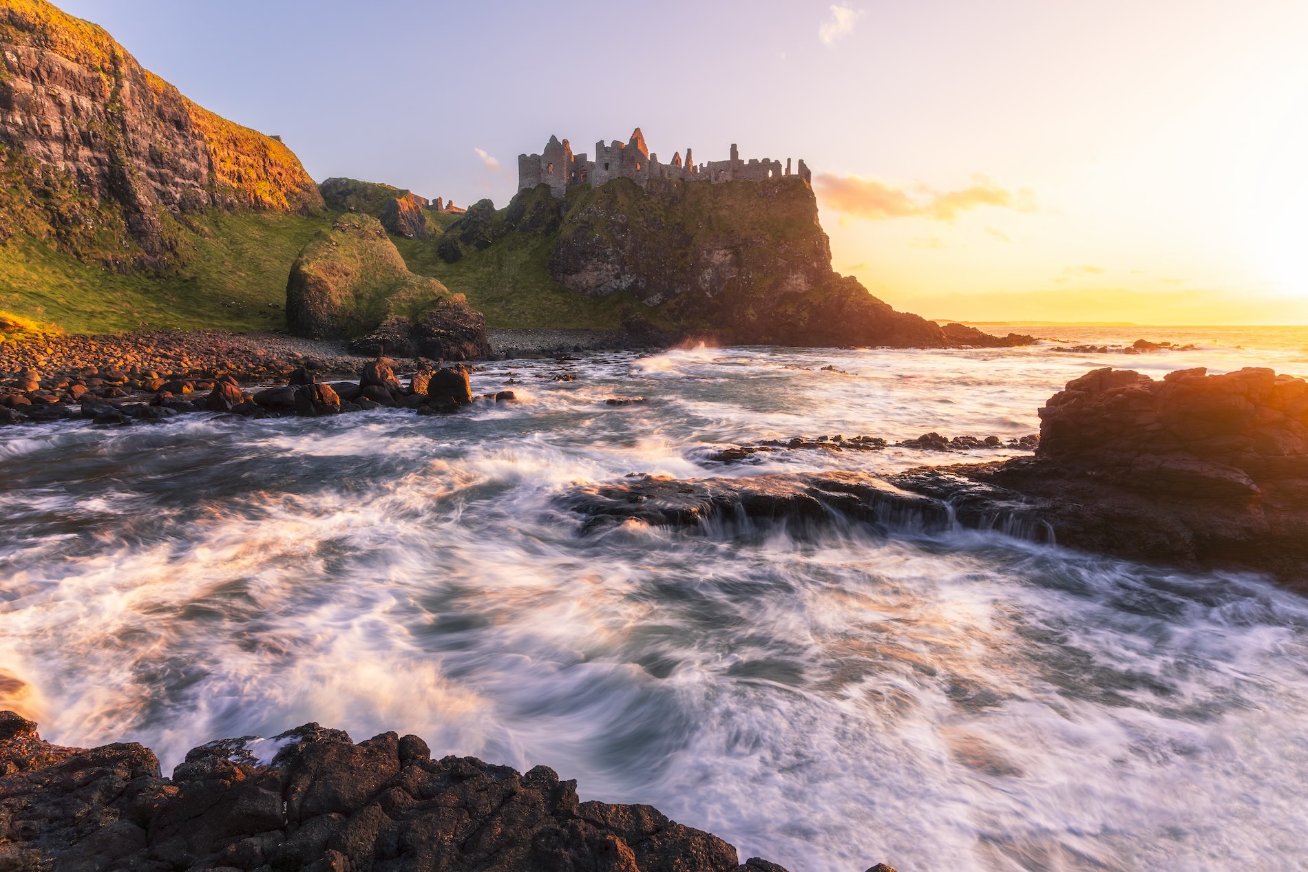 Waves surge against black rocks beneath the ruins of a cliffside castle at sunset.