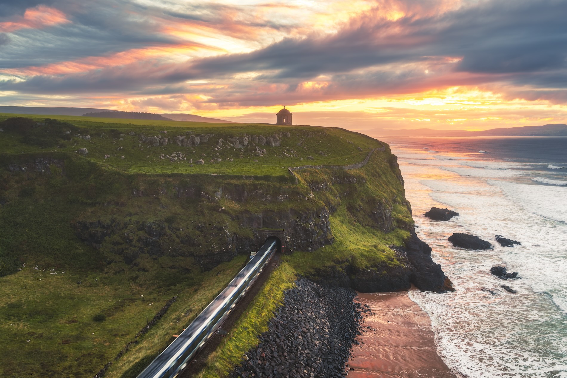 A clifftop temple at sunset overlooks waves crashing below and a train entering from a tunnel.