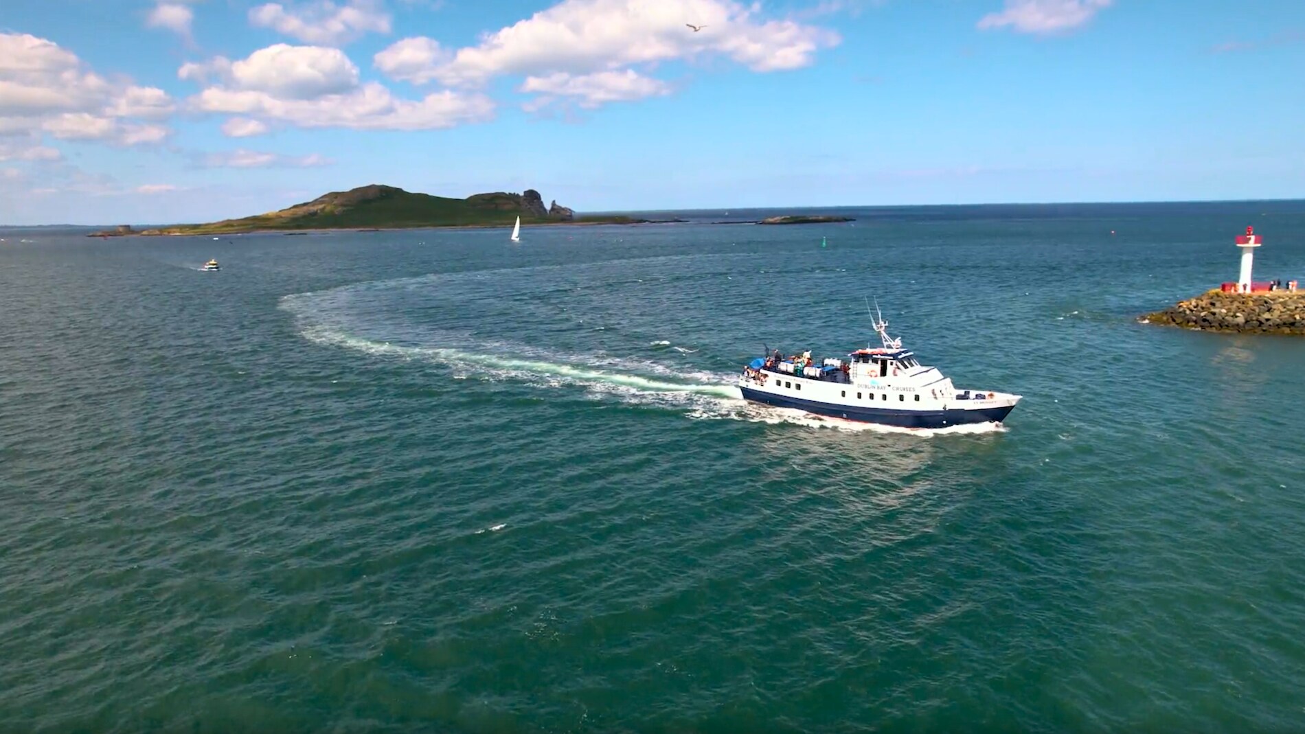 A sightseeing boat glides across the waters near a small lighthouse, with Ireland’s Eye in the distance.