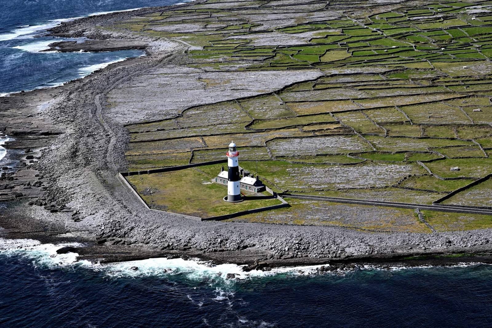 Black and white lighthouse surrounded by stone-walled green fields and a rocky coastline.