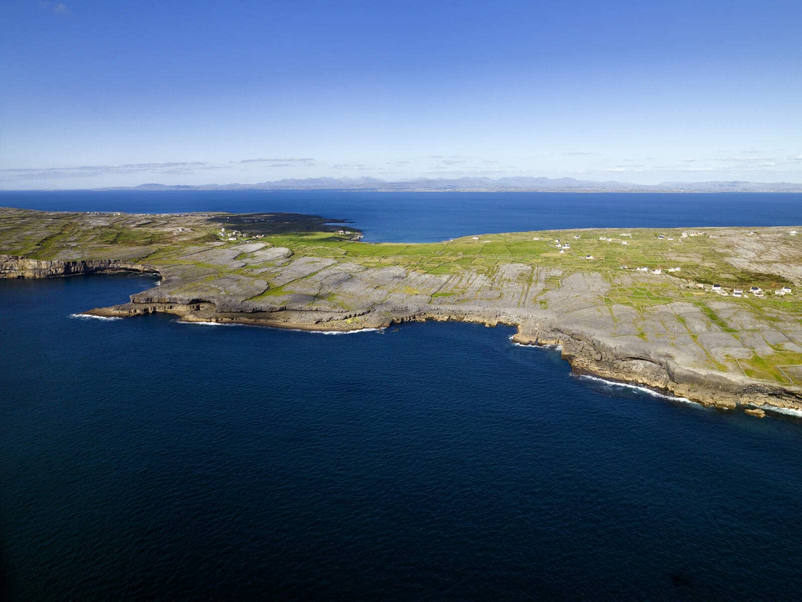 Rocky shoreline and patchwork fields of Inis Mór stretch toward the deep blue Atlantic Ocean under clear skies.