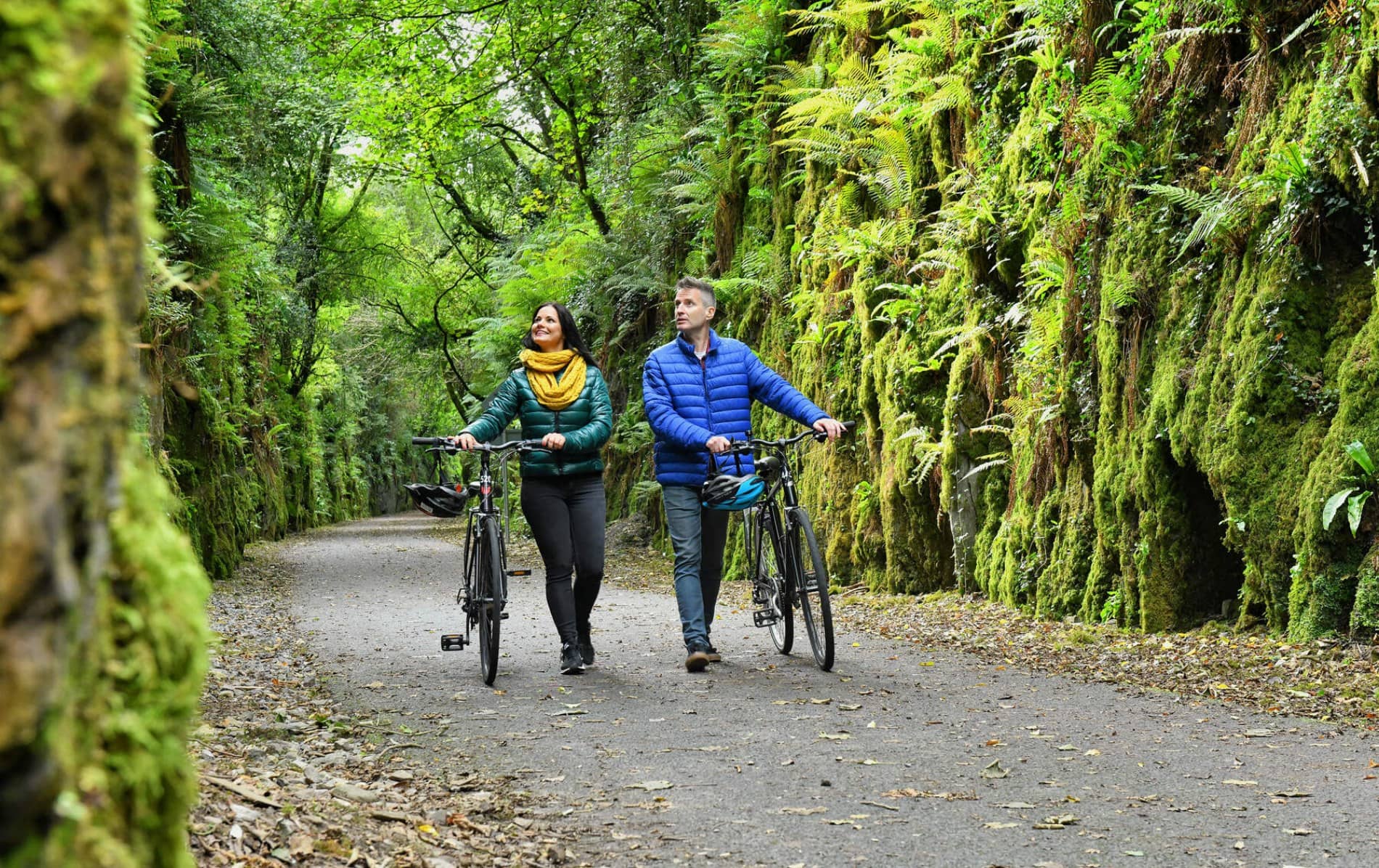 Two cyclists walking their bikes through a green, moss-lined path on the Waterford Greenway in County Waterford.