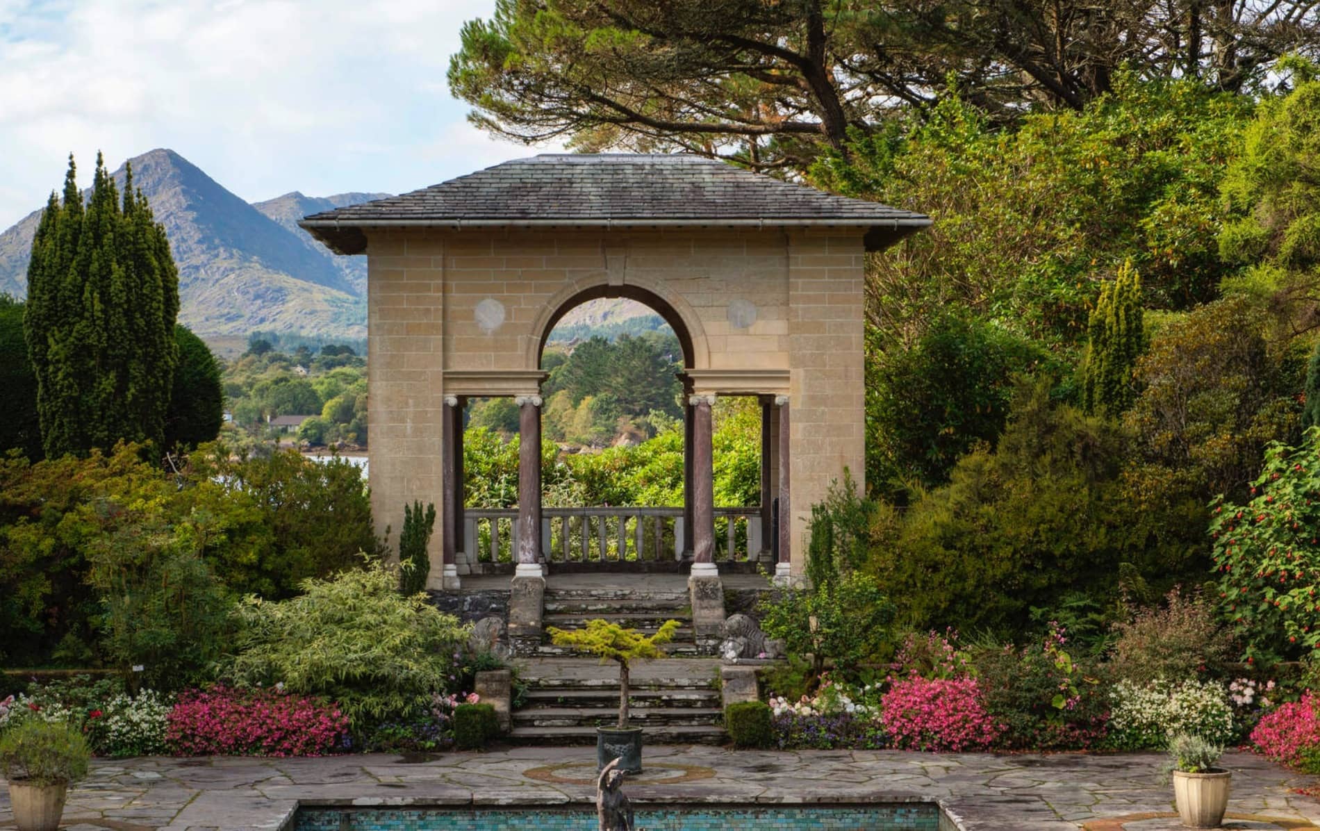 Un jardín formal en la isla de Garnish, Cork, con un elegante arco de piedra, macizos de flores cuidados y montañas al fondo.