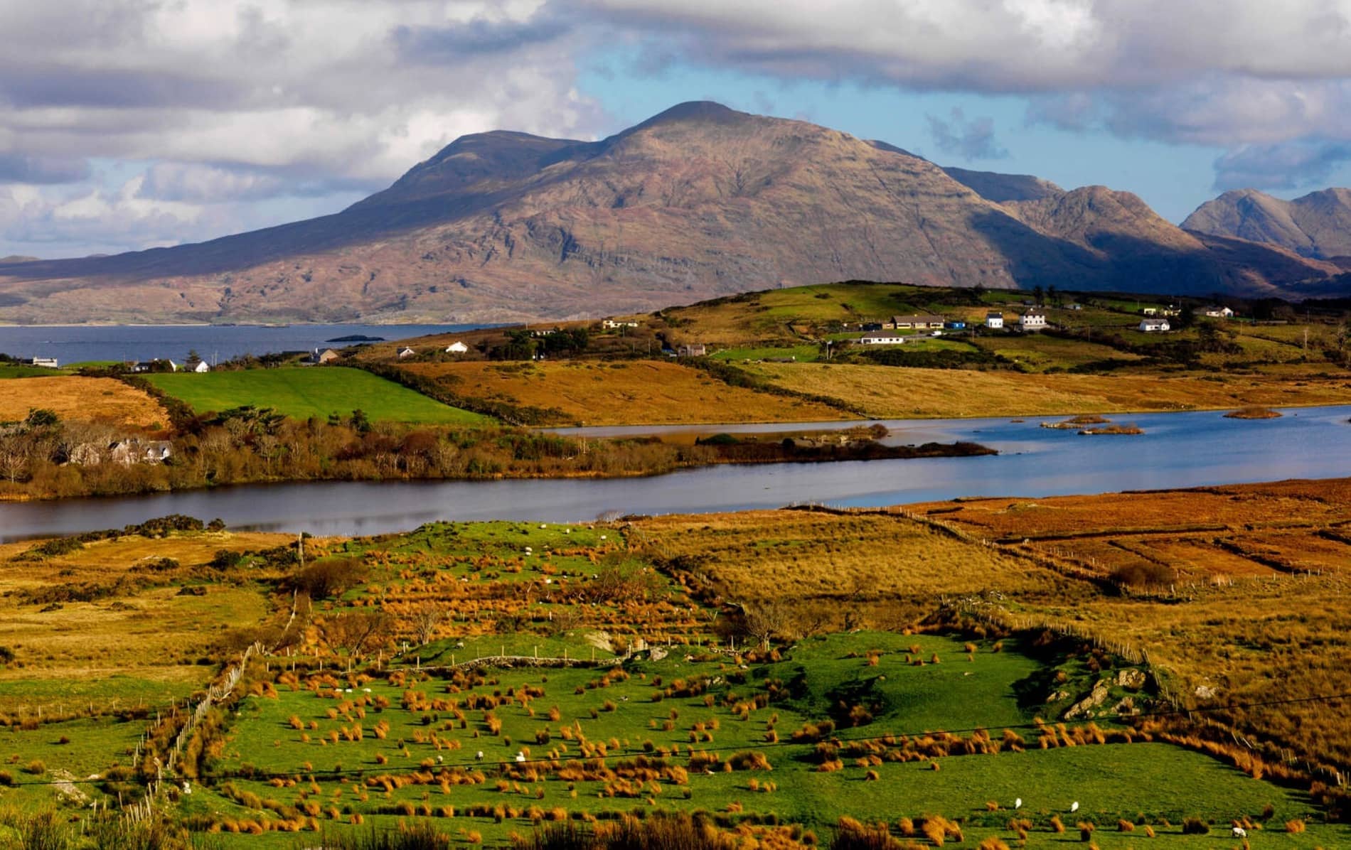 Un mosaico de campos verdes y dorados en el valle de Uggool, Mayo, con montañas elevándose detrás y un lago que refleja el paisaje.
