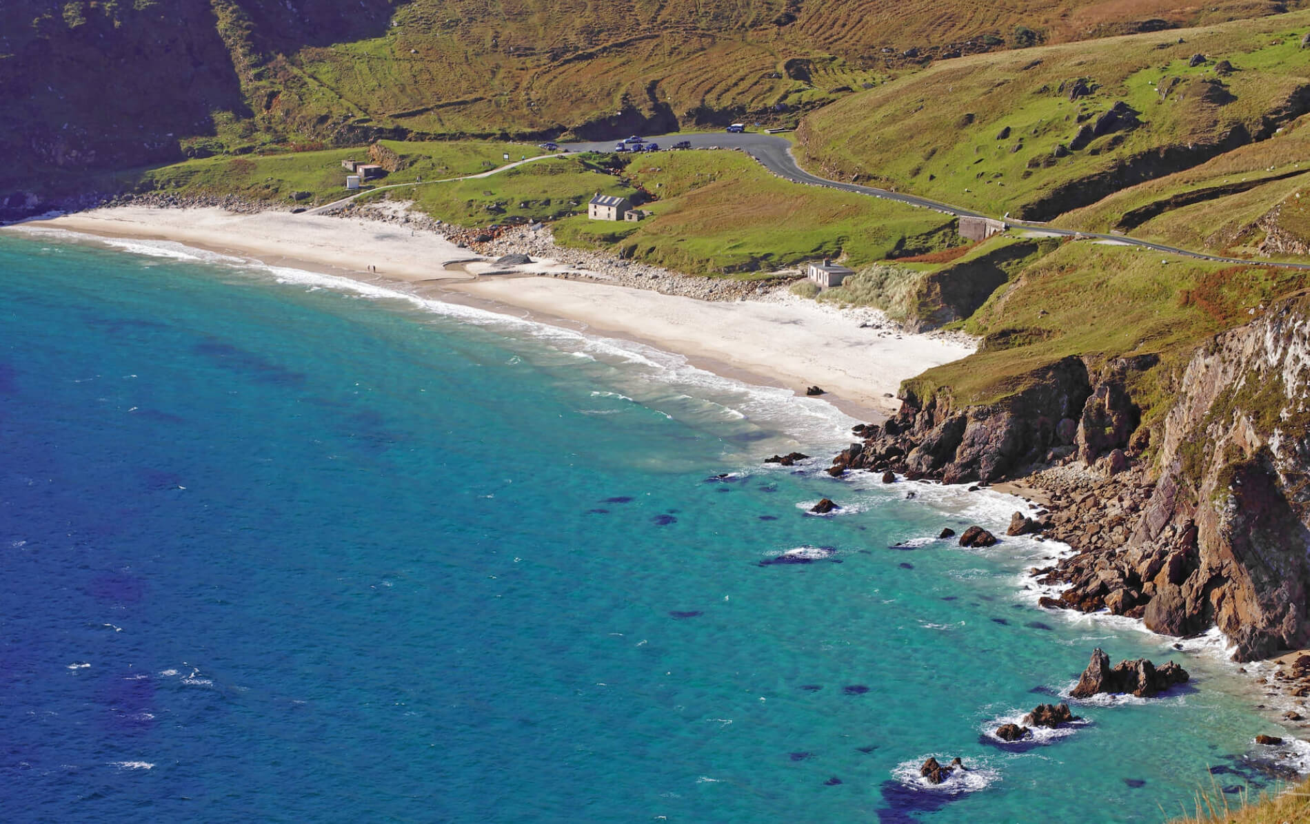 A curving stretch of white sand and turquoise water lies beneath rugged cliffs and grassy hills.