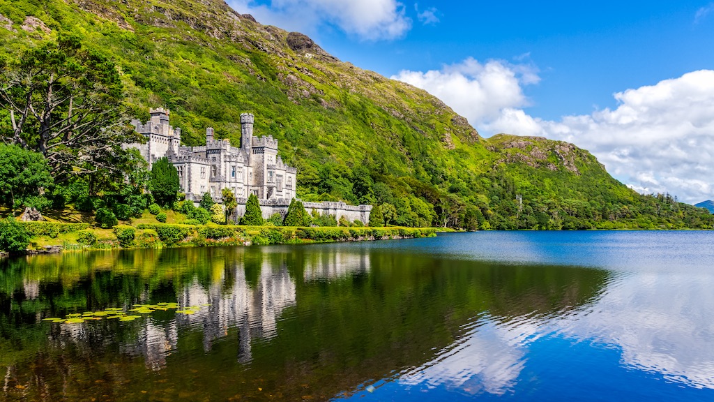 Kylemore Abbey mirrored in a glassy lake, set against forested hills and a blue sky.