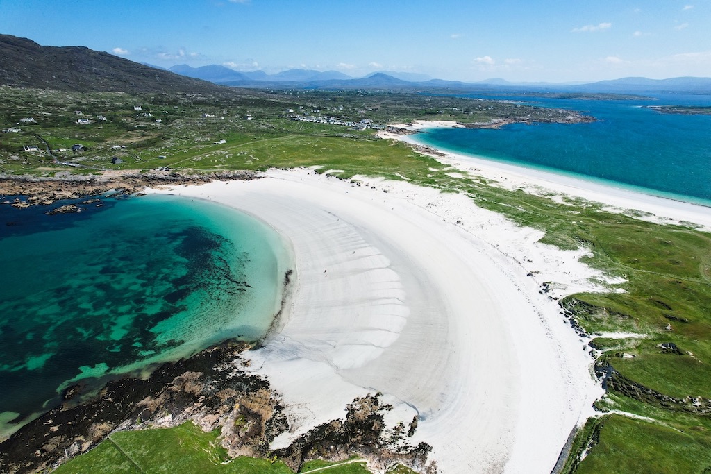 Dog’s Bay’s white sand curves around vivid turquoise water near Roundstone, with green fields and scattered houses inland.