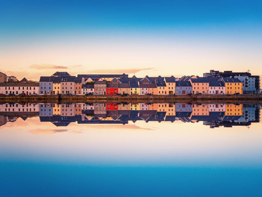 Row of colourful buildings reflecting on calm water during sunset along the waterfront in Galway.