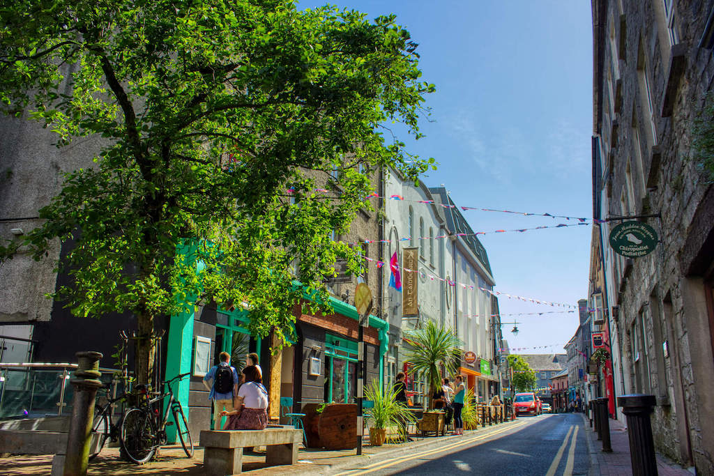 Tree-lined street in Galway city with hanging bunting, shopfronts, and people walking or sitting outside cafés.