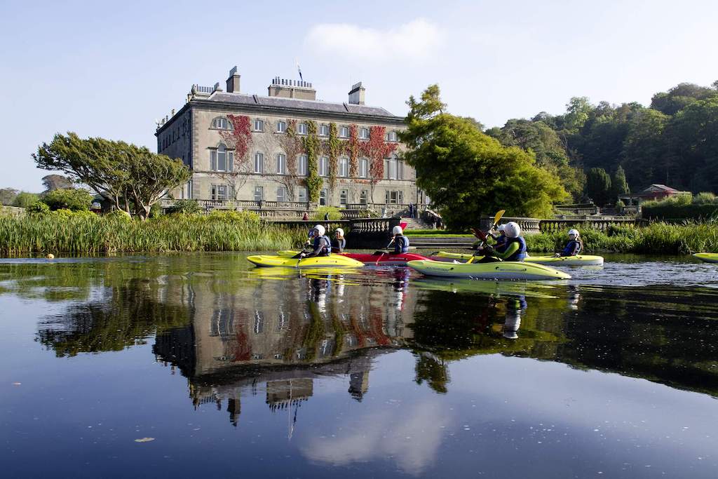 Group of people kayaking on a calm lake beside Westport House, with lush greenery in the background.