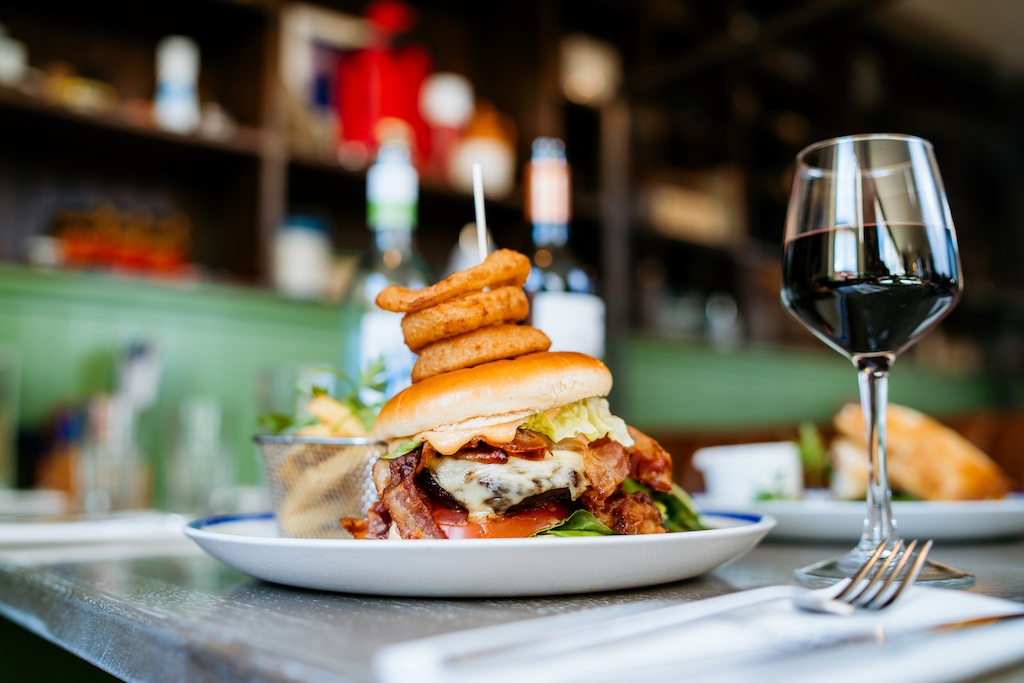 A stacked bacon cheeseburger topped with onion rings beside fries and a glass of red wine in Cobbler's Bay, County Mayo.