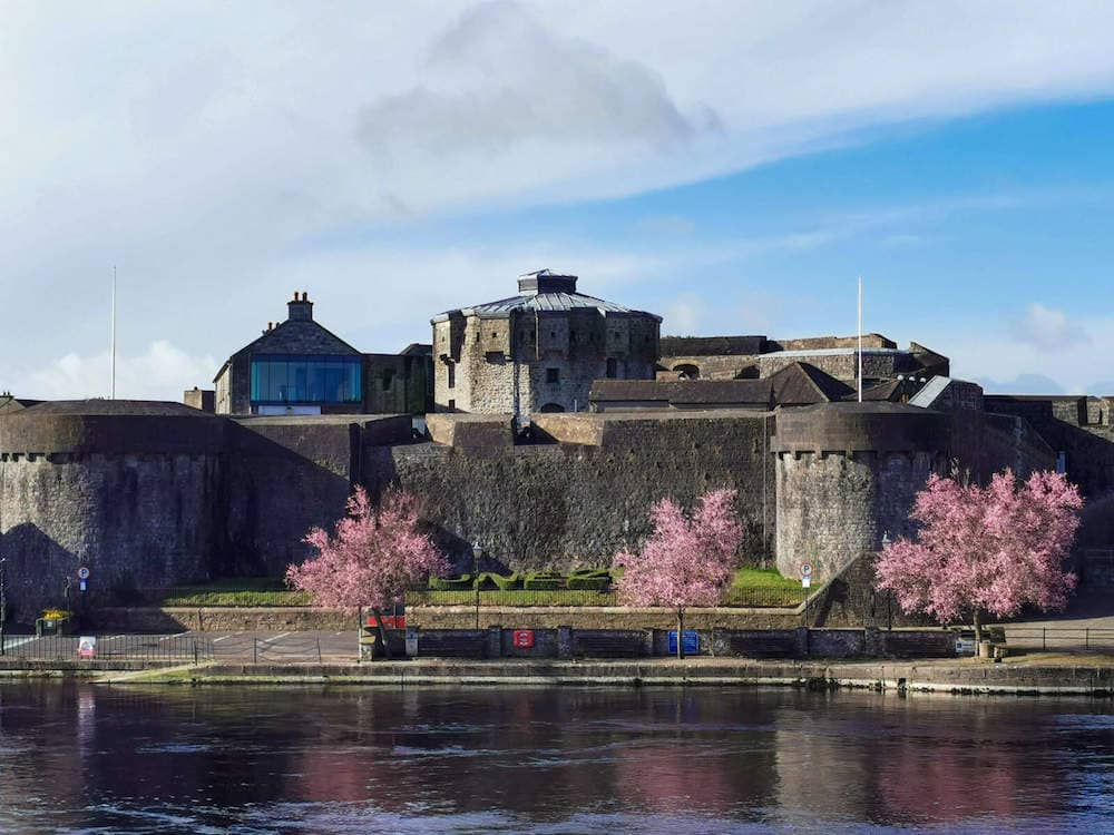 13th-century Athlone Castle on the riverbank, framed by pink cherry blossom trees.