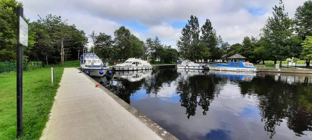 Boats docked at a peaceful marina surrounded by trees, viewed from a concrete path in Drumod.