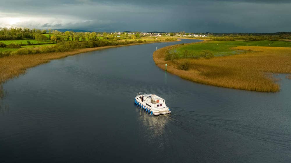 A cruiser travels through a winding river channel near Lough Key, bordered by reeds and green fields.