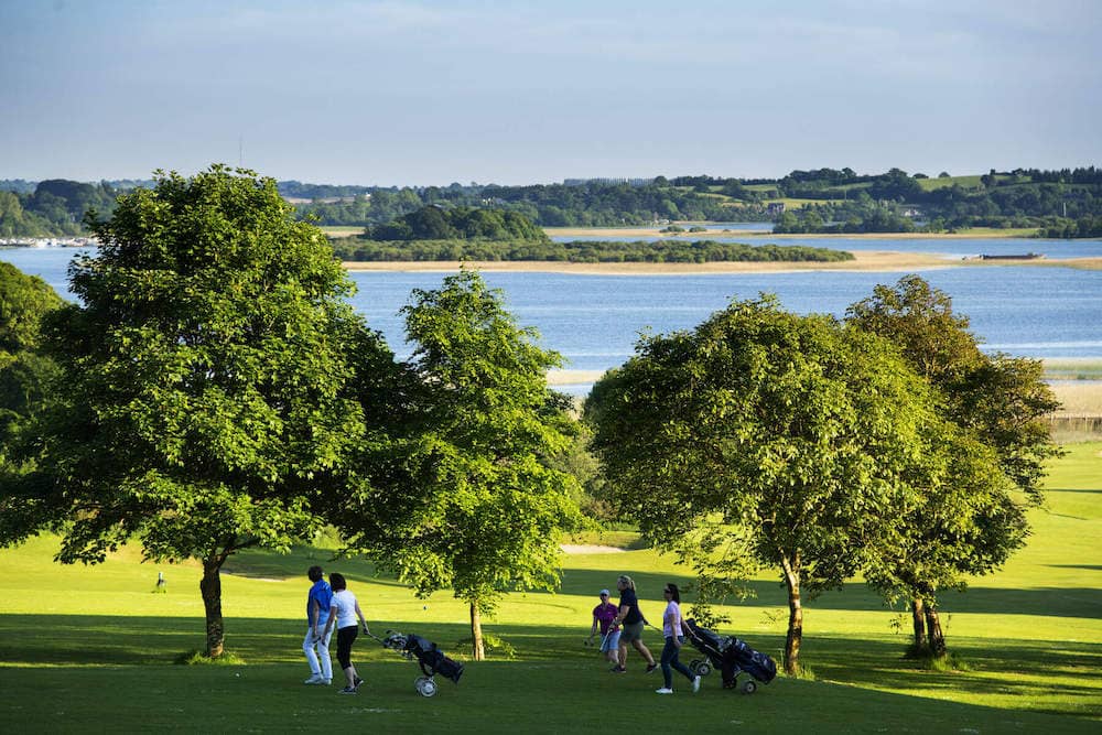 People walk with golf trolleys across green lawns at Glasson Lake House Hotel with lake views beyond.