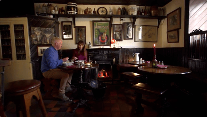Two people dining beside a lit hearth in the cosy, memorabilia-filled interior of Grogan’s pub in Glasson.