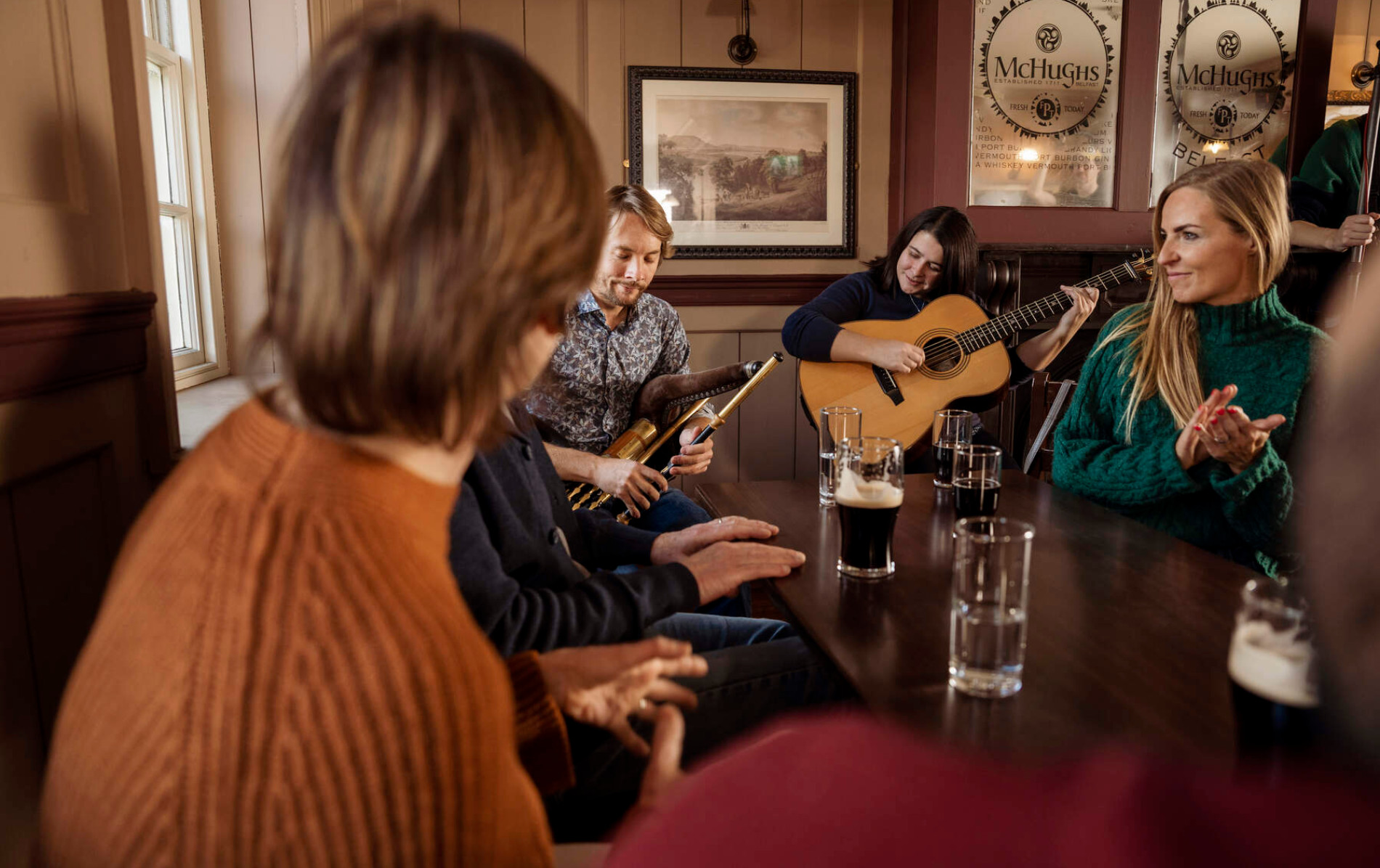 Musicisti e locali si godono una tradizionale sessione irlandese in un pub di Belfast, con risate e pinte condivise intorno al tavolo.