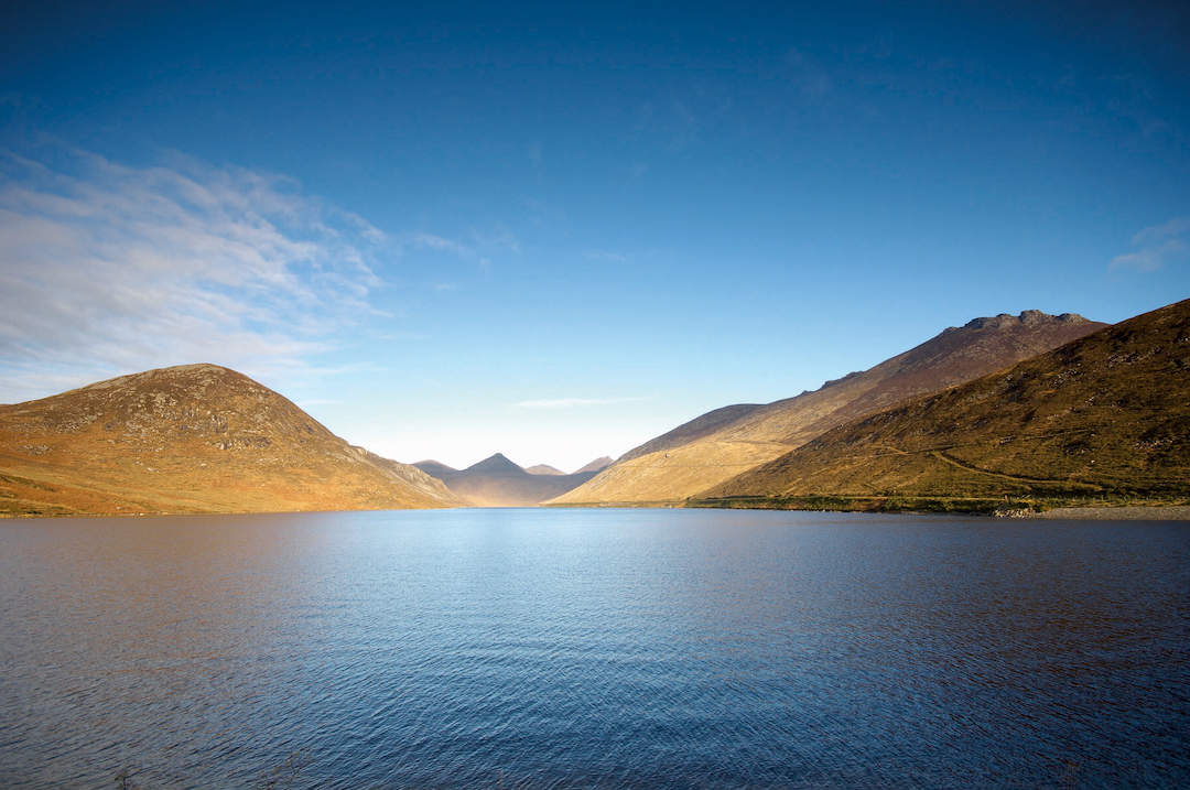 Ben Crom Reservoir nestled between rugged peaks of the Mourne Mountains under a clear blue sky.