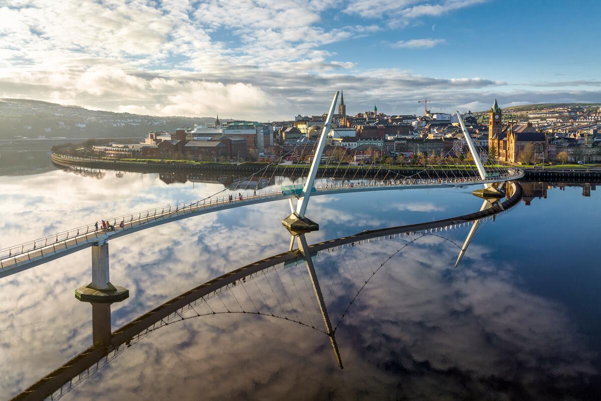De Peace Bridge weerspiegeld in de rivier de Foyle, met op de achtergrond de skyline van Derry~Londonderry.