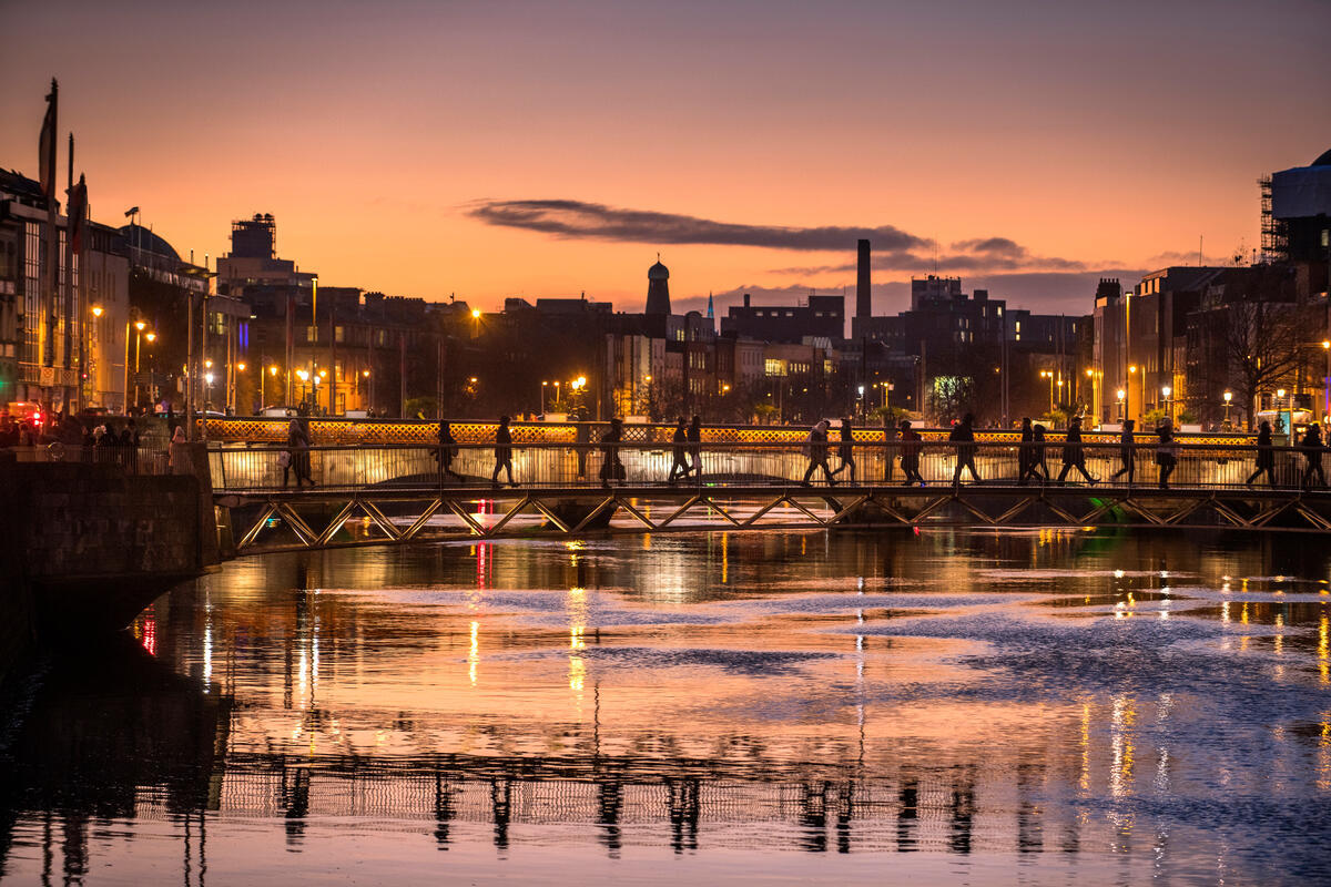 People crossing a bridge in Dublin at dusk with reflections on the River Liffey.