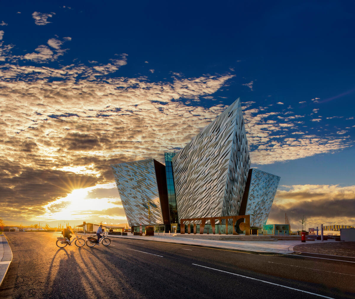 Titanic Belfast bei Sonnenuntergang: Radfahrer fahren vorbei und goldenes Licht liegt über dem Hafen von Belfast.