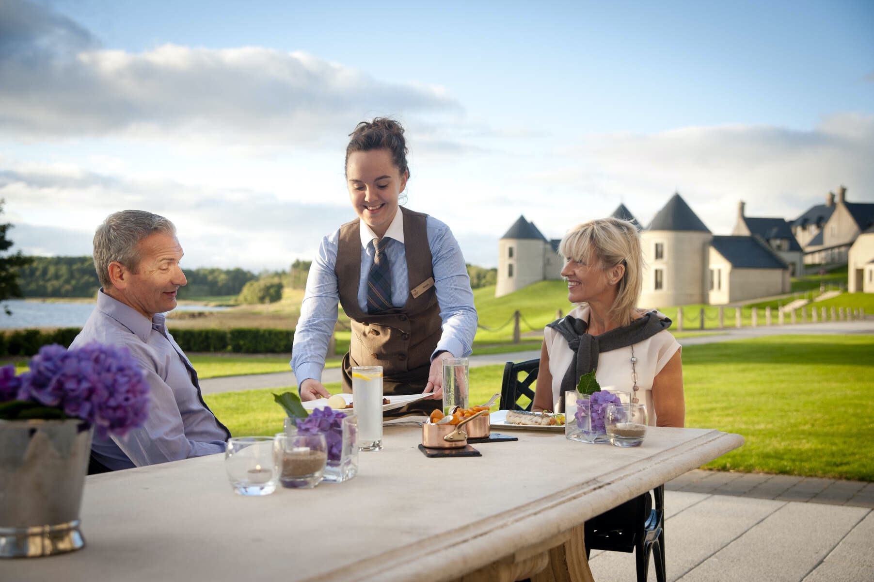 Waitress serving food to a couple dining outdoors at Lough Erne Resort with landscaped lawns, cottages, and lake views.