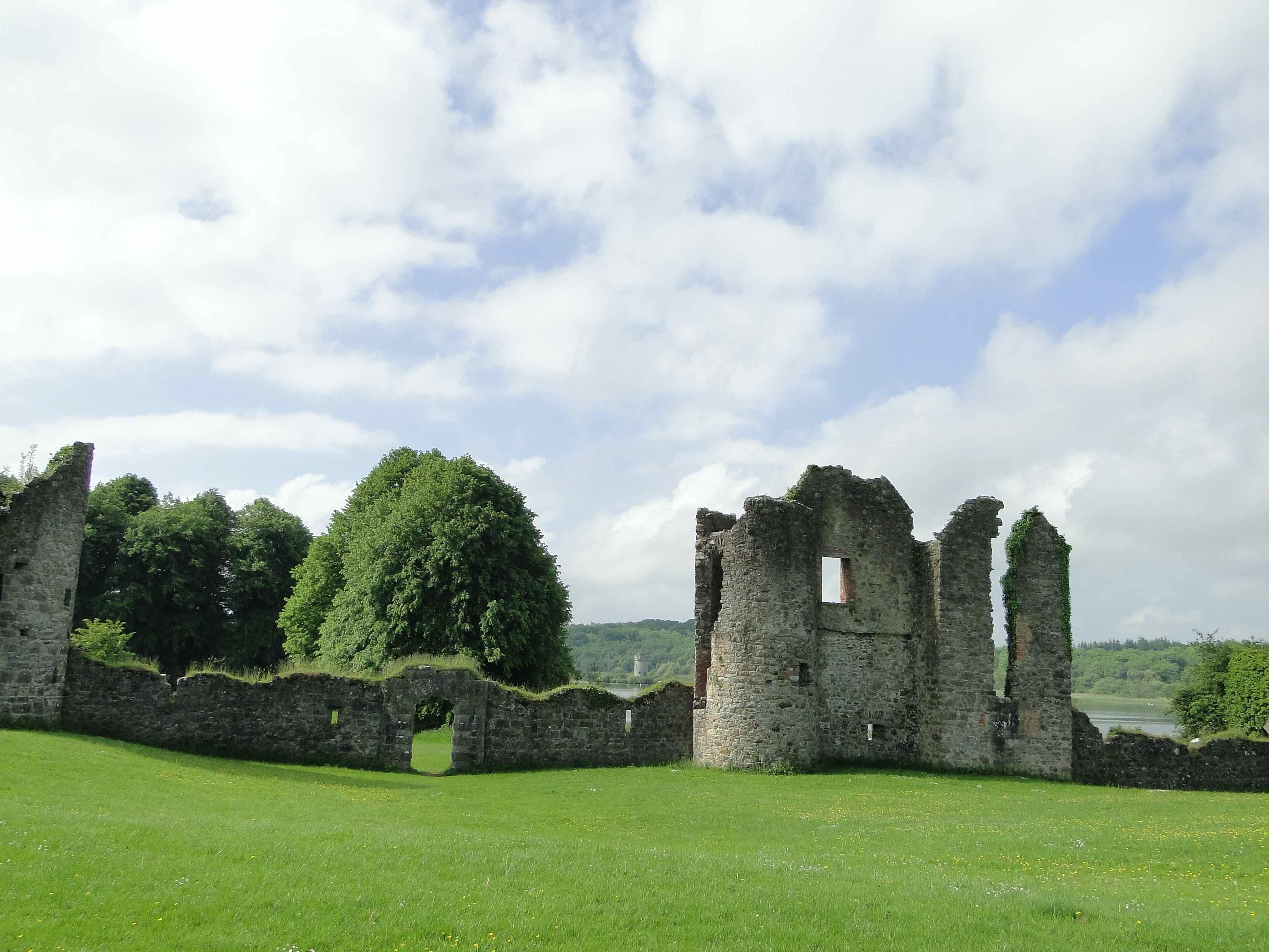 Stone ruins of Crom Estate framed by green lawns, leafy trees, and glimpses of Upper Lough Erne in the distance.
