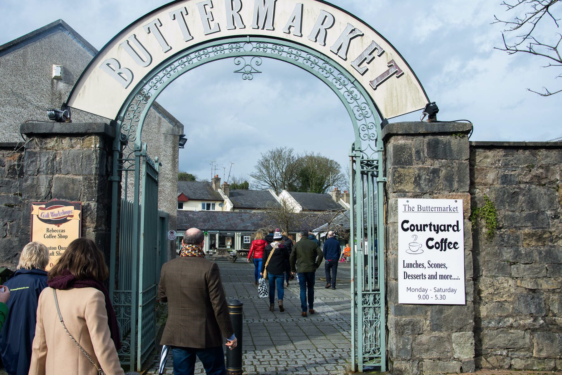Visitors walking through arched entrance of The Buttermarket courtyard with shops, coffee signs, and stone walls in view.